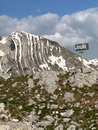 The image shows a view in Boričje, Montenegro, featuring a snow-capped mountain with a sign indicating "NP Durmitor" (National Park Durmitor). The mountain has distinct layered rock formations with patches of snow. The foreground consists of rocky terrain interspersed with patches of dry grass. The sky exhibits a mix of blue and white clouds.