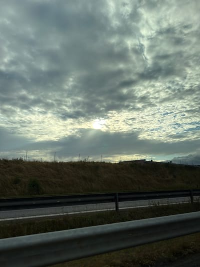 The image shows a cloudy sky with sunlight breaking through in the upper central part of the frame. Below the sky, there is a grassy embankment with a chain-link fence running horizontally across it. Beyond the fence, a building or structure is visible on the horizon. In the foreground, there is a road with guardrails. The scene appears to be along a highway in Tofta, Sweden, based on the general landscape and the presence of typical Swedish roadside infrastructure. There are no people or animals visible in the image.