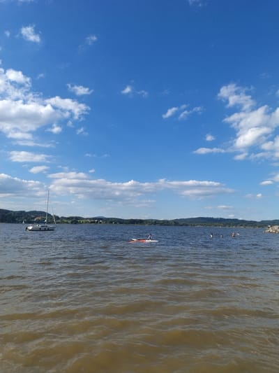 The image shows a wide shot of a lake on a sunny day with scattered white clouds against a bright blue sky. In the middle ground, a white sailboat with a single mast is docked. Further out on the water, several people are swimming and one person is on a paddleboard. The shoreline in the background features a treeline with some scattered buildings and hills. The water in the foreground is a murky brown color with small waves. The overall scene depicts a recreational activity at a lake.