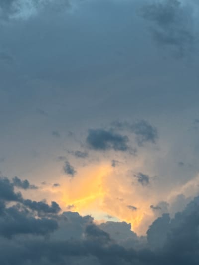 The image shows a cloudy sky with dramatic lighting. A patch of bright orange and yellow light shines through the dark clouds, creating a striking contrast. The clouds are dark and heavy, suggesting an impending storm or the aftermath of one. The sky is predominantly a deep blue-gray, with lighter shades of blue visible in the upper portion of the image. There are no people, structures, or geographical landmarks visible in the image that would indicate its specific location within the city of Gorno-Altaysk, Russia. The scene is entirely focused on the natural phenomenon of the sky and clouds.