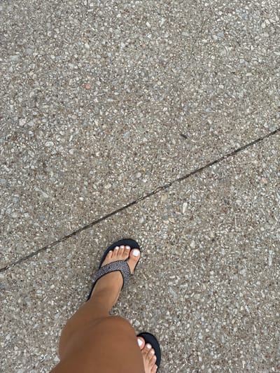 A pair of feet wearing black flip-flops with white-painted toenails stand on a cracked concrete surface. The foot on the left is closer to the camera, with its toes and a portion of the leg visible. The foot on the right is partially visible at the bottom edge of the frame. The concrete is speckled with small stones and pebbles, and a dark, thin crack runs diagonally across the surface. The scene appears to be outdoors, on a sidewalk or pathway, in Balch Springs, United States of America.