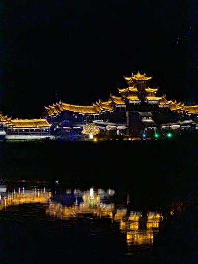 The image depicts a nighttime scene of a waterfront area in Zhuoshui, China. The photo is rotated 90 degrees clockwise.

A structure with traditional Chinese architectural elements is visible on the left side, illuminated with golden lights. There is a walkway and buildings with green and white lighting. The right side of the image shows a reflection of the waterfront area in the water. The water reflections appear blurred due to waves or movement. There are no people visible.
