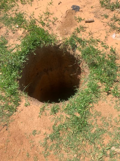 An aerial view of a large, circular hole dug into reddish-brown soil. The hole is deep and dark, with rough, earthy walls. Green grass grows in clumps around the rim of the hole and sparsely across the surrounding soil. The ground appears dry and somewhat uneven. To the upper right of the hole, a few small, light-colored rocks are visible, as well as a piece of dark, possibly plastic, debris. Further to the right and slightly above the hole, a cylindrical object with horizontal ridges, resembling a spool or cap, lies on the ground. The scene is outdoors, with natural lighting casting shadows. The location is identified as Buga da Gindi, Nigeria.