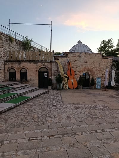 The image depicts the entrance of a jazz museum in Gradsko opština Crveni Krst, Serbia, during twilight. The building is constructed of stone with a domed roof. Prominently displayed outside are oversized, stylized representations of a saxophone and a double bass. A sign indicates "JAZZ MUSEUM" above the entrance, which has two arched windows to its left. Steps leading up to the entrance are covered with green turf and feature repeated text that reads "NISVILLE JAZZ MUSEUM". The surrounding area is paved with cobblestones. In the background, a stone wall and trees are visible against a pale sky with a sliver of moon and soft clouds. There are no people visible in the immediate vicinity of the entrance.