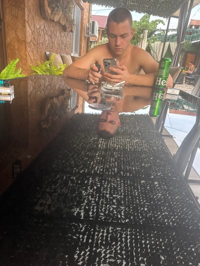 A young, shirtless man with short brown hair is seated at a reflective, dark table outdoors. He is holding a smartphone in his left hand and an object resembling a lighter in his right hand. To his right, a green can with the "Heineken" logo is visible. In front of him, a clear glass with a cigarette in an ashtray sits on the table. The table's surface reflects the man, the can, and the surrounding environment. In the background, a stone-textured wall with decorative plants and an air conditioning unit is visible. Further back, there are couches and greenery. The scene appears to be a patio or outdoor seating area, possibly in a residential or recreational setting. The context of Chiprovtsi, Bulgaria, is not visually discernible from the image itself.