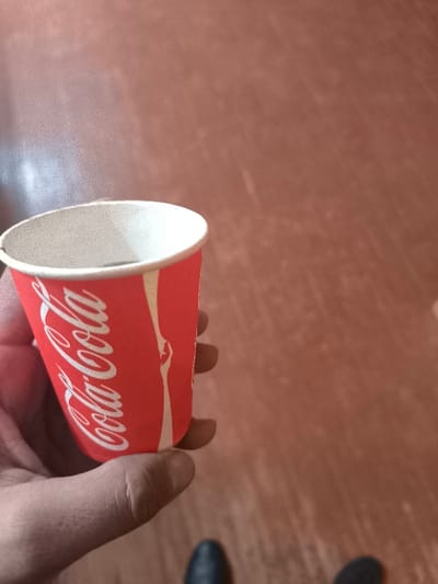 The image depicts a close-up view of a human hand holding a red and white paper cup, branded with "Coca-Cola" in white script. The cup contains a dark liquid, visible near the white rim. The hand, exhibiting visible skin texture, grasps the cup from the left side of the frame, with the thumb, index, and middle fingers primarily visible. The background consists of a uniform reddish-brown surface, likely a floor, with subtle variations in tone. At the bottom edge of the frame, the tops of two dark shoes are partially visible, suggesting the individual holding the cup is standing and looking downwards. The scene is located in the city of Pakhtakor, Uzbekistan.