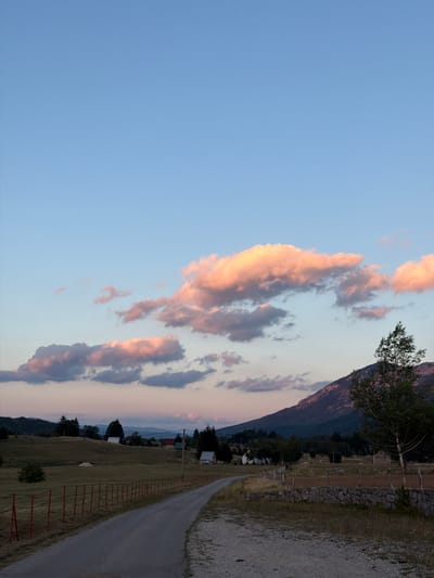 The image depicts a rural landscape in Donja Brezna, Montenegro, during twilight. A paved road curves into the distance, bordered by a dry-stone wall on the right and a fence with red posts on the left. Rolling hills, dotted with scattered houses and trees, stretch towards distant mountains. The sky is a gradient of soft blues and pinks, with illuminated clouds reflecting the sunset. A lone tree stands on the right side of the frame, silhouetted against the mountains. The scene is tranquil and devoid of human presence.