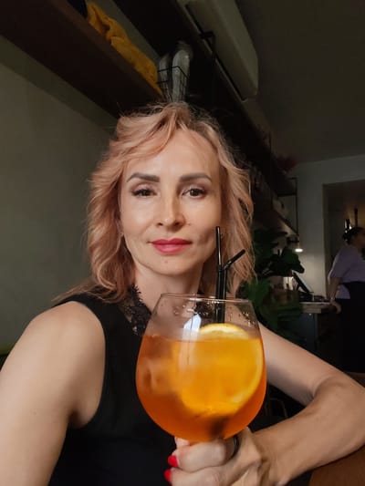 The image shows a medium shot of a woman with light pink hair holding a glass of an orange cocktail with a slice of lemon in a dimly lit indoor setting, possibly a cafe or restaurant. She is wearing a black sleeveless top with lace detailing around the neckline. Her right hand is holding the glass, and she is wearing a ring on her left hand. In the background, shelves with items are visible, as well as some plants and other patrons. The overall lighting suggests an evening or indoor environment. There is no visible indication that this scene is located in Selivanovo, Russia.