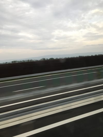 The image depicts a view from a moving vehicle on a highway in Adjud, Romania, under an overcast sky. The upper half of the frame is dominated by a light grey, cloudy sky with subtle brighter areas. Below the sky, distant, hazy hills and a thin line of deciduous trees form the horizon. In the midground, a dark, elevated earthen embankment or a freshly tilled field runs parallel to the highway. The lower half of the image features a multi-lane asphalt highway, characterized by solid and dashed white lane markings. A metallic silver guardrail separates the immediate foreground lanes from a narrow strip of green vegetation, which in turn borders the dark earthen area. Motion blur is evident, particularly in the highway lanes and guardrail, indicating the photograph was taken from a vehicle in motion. No individuals, other vehicles, or specific actions are discernible.