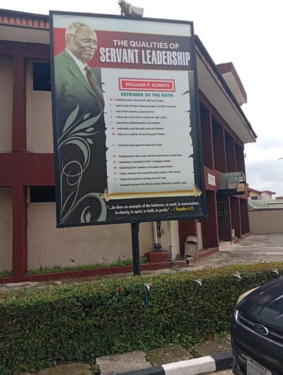 This image captures a detailed view of a billboard featuring "The Qualities of Servant Leadership" and the image of a man, identified as William F. Kumuyi. The billboard is positioned outdoors, in front of a building with a maroon and beige facade, typical of architecture found in cities like Eliozu, Nigeria. The billboard itself is large and prominently displays text and graphics related to leadership qualities, listed with alphabetical acronyms. The surrounding environment includes lush green hedges, a black car, and concrete pathways. The scene appears to be a sunny day, casting shadows on the ground. The building behind the billboard has windows and doors, suggesting it's a public or commercial space. The overall impression is one of a community or religious institution displaying inspirational messages.