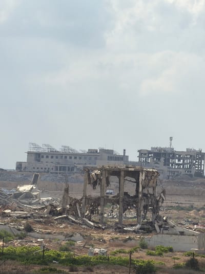The image depicts a scene of widespread destruction. Several partially or completely destroyed buildings dominate the landscape, with concrete debris scattered across the ground. The buildings appear to be multi-story structures, some with exposed rebar and collapsed floors. The architectural style suggests they were likely residential or commercial buildings.

In the foreground, closer to the viewer, a heavily damaged concrete structure stands, revealing its internal framework and the extent of the structural failure. Further in the background, more buildings are visible, some appearing to be in a state of partial collapse with damaged facades and missing sections. The overall impression is one of a recently devastated urban or semi-urban area.

The environment around the destroyed buildings is dry and arid, with sparse vegetation in some areas. Patches of green foliage, possibly agricultural land or trees, can be seen in the mid-ground and background, contrasting with the gray rubble. The sky is overcast and hazy, suggesting a dull or possibly humid day. There are no visible people in the image, focusing entirely on the aftermath of destruction.

The scene evokes a sense of desolation and loss. The destruction is extensive and appears to be the result of significant force, possibly from conflict or a natural disaster. The location context of Az-Zahra, Palestinian Territories, points towards the possibility of this being a consequence of military action. No text is visible in the image.