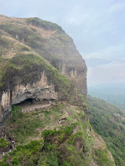 The image depicts a steep mountain face in the city of Planes de Renderos, El Salvador. A paved path, lined with what appear to be white, concrete markers, runs along the side of the mountain, leading to a cave entrance. People are seen walking along the path and on steps at the bottom of the image. Vegetation covers portions of the mountain, with trees and shrubbery visible on the right side. The sky is overcast, and a valley with more vegetation and structures is visible in the background. The mountain face itself is a mix of rock and greenery.