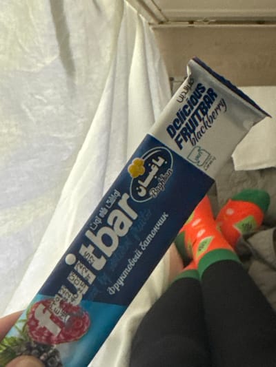 A close-up image shows the lower body of a person, positioned in what appears to be an indoor setting with light-colored fabric. The person's left hand holds a partially unwrapped "Delicious FRUITBAR blackberry" produced by "Baghan," with text in English, Arabic, and Cyrillic script ("фруктовый батончик"). The packaging is predominantly blue and white, featuring an image of raspberries and blackberries. The person is wearing black leggings or pants and vibrant orange socks with green cuffs and a watermelon pattern. The background consists of draped white or light-colored bedding or fabric, and an indistinct light-brown surface above. The precise geographical location within совхоза "Воронежский", Russia, cannot be determined from the visual information presented.
