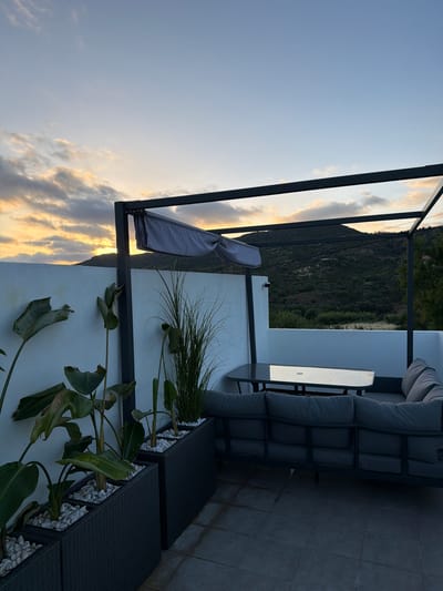 The image depicts a rooftop terrace at dusk, overlooking a verdant, hilly landscape. The sky is a soft gradient of blue and orange, with scattered clouds catching the last rays of the sun. A modern outdoor seating area is arranged under a dark metal pergola with a grey fabric canopy. The seating consists of a large, U-shaped sectional sofa in a dark grey fabric, facing a dark-framed table with a glass top. Adjacent to the seating area, several large planters contain lush green foliage, with one tall, feathery grass-like plant and broad-leafed plants. The terrace floor is paved with square grey tiles. The white wall bordering the terrace is visible, with the sun setting behind the hills in the background. The scene suggests a peaceful, luxurious outdoor space in a natural setting, likely a residential property or a boutique hotel in the Loulé region of Portugal.
