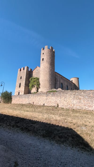 The image shows a stone castle with a tall, round tower and several crenelated rectangular towers, situated in Chambles, France. A thick stone wall surrounds the castle, and a dry, grassy hill leads up to it. A gravel path is visible in the foreground, casting a long shadow. A lamppost and some bushes are present to the left of the castle. The sky is clear and blue.