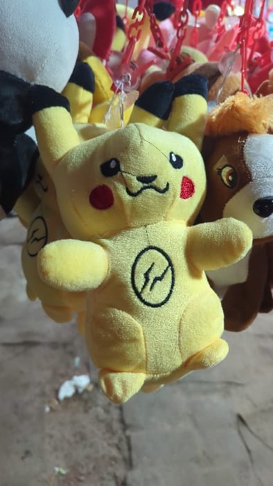 A close-up shows a cluster of plush toys. A yellow Pikachu toy with black ears and a red cheek is prominently featured, displaying a stitched lightning bolt symbol on its back. Above Pikachu, a brown animal toy is partially visible. Red plastic chains are attached to the toys. A black and white panda toy is also partially visible at the bottom of the image. The background appears to be a blurry wall.