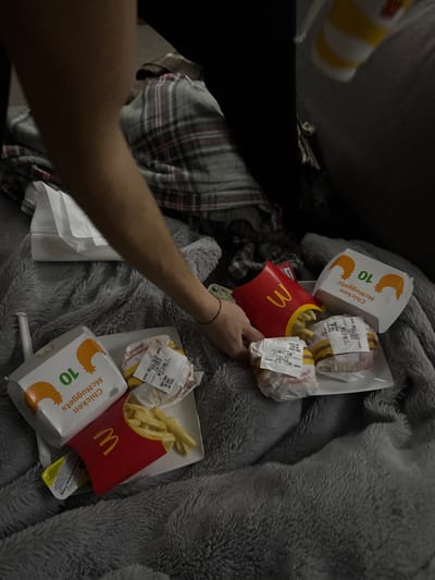 A person's arm reaches towards a pile of McDonald's food on a gray blanket. The food includes two boxes of 10-piece Chicken McNuggets, two red French fry containers, two burgers wrapped in paper with receipts attached, and two white plates. One plate holds fries and a burger, while the other appears to hold two burgers and fries. There is a small packet of sauce near one of the plates. A plaid shirt is visible in the background.