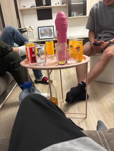 The image shows an indoor scene, with several people seated around a small, pink, two-tiered serving cart filled with various glasses and bottles.
People: The image captures portions of at least three individuals. One person wears a grey t-shirt and shorts, holding a black object in their hand. Another is wearing black sweatpants. A third person is wearing jeans and an olive green jacket.
Objects: A pink serving cart holds several items, including a pink dildo, glasses with the logos of Coca-Cola and Lipton Ice Tea, and what appears to be a large bottle of water. A framed picture hangs on the wall.
Environment: The scene appears to be inside a home, with visible furniture and décor. A wooden floor is visible, and a cabinet is positioned against the wall.
The location in the city of Graf Ignatievo, Bulgaria is unknown.