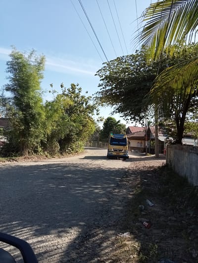 The image depicts a dusty, unpaved road in a rural or suburban area, likely Buketglumpang, Indonesia. The road is flanked by dense green foliage, including bamboo on the left and palm trees on the right. In the middle ground, a yellow Mitsubishi Fuso truck is driving towards the viewer, kicking up dust. Several houses with reddish-brown tile roofs are visible behind the trees. The sky is clear blue with a few thin wisps of clouds. The foreground shows the gravelly road surface with shadows from trees cast across it. Discarded plastic bottles and wrappers are scattered on the ground on the right side of the road. The scene suggests a warm, sunny day.
