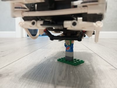 A low-angle, close-up shot captures a LEGO minifigure standing on a green LEGO base plate. The minifigure wears a blue shirt with a red and white emblem on the chest and a green pixelated head. It is positioned directly beneath the chassis of a white and black LEGO vehicle, which appears to be a car. The underside of the LEGO vehicle is detailed with various structural components, including white arms and black support beams.
The setting is an indoor environment, with a light-colored wooden floor providing the surface. The floorboards are laid horizontally in the frame and show a subtle wood grain pattern. In the background, the out-of-focus shapes of a white door frame and a wall are visible, suggesting a domestic or indoor space. The lighting appears to be artificial, casting soft shadows from the LEGO elements onto the floor.
The image is static, with no discernible activity or event taking place beyond the posed arrangement of the LEGOs. The focus is on the miniature scale of the minifigure in relation to the larger LEGO vehicle, creating a playful and somewhat whimsical scene. There is no visible text within the frame. The overall mood is neutral and observational.