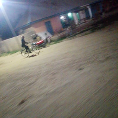 The image captures a person riding a bicycle at night in a motion blur. The rider, dressed in dark clothing, is seated on a silver bicycle with a bright red object, possibly a bag or cargo, attached to the rear rack. The bicycle has two wheels, with the front wheel appearing slightly larger than the rear. The rider's left hand is on the handlebars. The scene appears to be outdoors, with a building in the background that has illuminated windows and a dark doorway. The ground is a light-colored, textured surface, possibly a dirt road or pathway. The overall lighting suggests it is nighttime, with a bright light source in the upper left quadrant of the image, possibly a streetlamp or headlights, casting a glare. The blur indicates movement of the camera or the subject. This scene is described as being in Wuro Allahamdu, Nigeria.