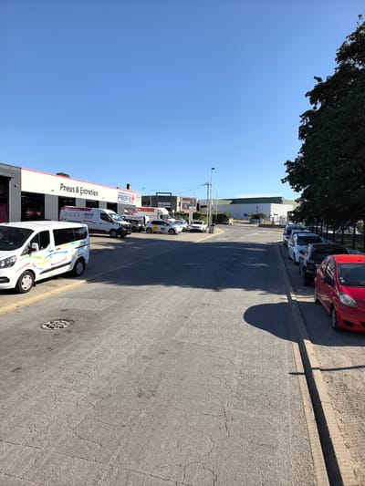 A wide, outdoor, eye-level shot captures a street scene in Saint-Herblain, France, under a clear blue sky. In the foreground, a cracked asphalt road stretches toward the right, with a manhole cover visible on the left. A curb runs along the right side of the road, with several parked cars lining it. The closest car is a red, small hatchback. Behind it are a dark sedan, a light blue sedan, and a darker blue sedan.
On the left side of the image, a white van with colorful decals is parked near a building that appears to be a business. This building has a light-colored facade with a pink stripe and the text "Pneus & Entretien" and "PROFIL". Adjacent to this, a white panel van with "PROFIL" branding is parked. Further back, other vehicles and buildings are visible, including a long, light-colored industrial building with a green roof in the distance. A lone street light stands on a pole to the left of the road. Dense green trees are visible on the far right of the frame, with their leaves casting shadows. The scene appears to be a commercial or industrial area.