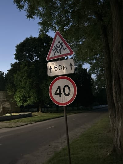 The image shows a road in the vicinity of Pariski village council, Belarus. Three traffic signs are mounted on a pole. The uppermost sign is a red-bordered triangle with a silhouette of children running, indicating a pedestrian crossing or school zone. Below it, a rectangular sign with a black arrow pointing upwards and the text "50M" signifies a distance of 50 meters. The lowest sign is a circular speed limit sign with a red border and the number "40" in black, indicating a maximum speed of 40 kilometers per hour. The signs are positioned along a paved road lined with grass and trees. The sky is clear and blue, suggesting it is daytime or early evening. The environment appears to be a rural or suburban setting.