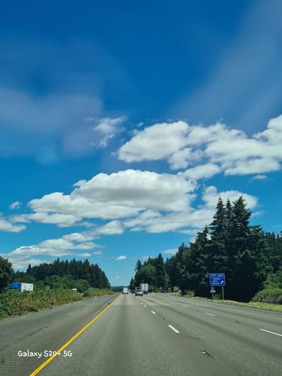 A wide-angle, slightly motion-blurred shot captures a highway in Aurora, United States, under a bright blue sky dotted with white, fluffy clouds. The road, marked with a yellow line, stretches into the distance, bordered by lush green trees on either side. Several vehicles, including trucks and cars, are visible on the highway. A blue sign indicating "REST AREA TRAVEL INFO CENTER" with an arrow pointing to the right is positioned on the right side of the road. The lower left corner of the image features the text "Galaxy S20+ 5G." The overall scene suggests a sunny day on a freeway, likely during daylight hours.