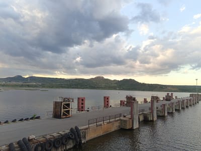 The image shows an old pier extending into the water. The pier is made of concrete and asphalt, with weathered metal structures and railings. There are tires along the edge of the pier. In the background, there is a hilly landscape under a cloudy sky. The image was taken in the city of Beloslav, Bulgaria.