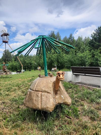 In the city of Zelenogorsk, Russia, on a grassy lawn under a cloudy sky, a whimsical metal sculpture of a turtle wearing oversized goggles sits prominently. Its shell is rough and tan, resembling a large rock, and a green, umbrella-like structure emerges from its back. To the left, another metal sculpture of a mosquito-like creature perches atop a tall, rusty pole. Further in the background, a metal sculpture of a person riding a bicycle is visible, along with other abstract metal artworks. A wooden park bench sits to the right of the turtle sculpture, positioned near a paved pathway that leads towards a dense forest bordering the area. Several people are scattered throughout the park, appearing to observe the sculptures and enjoy the outdoor space.