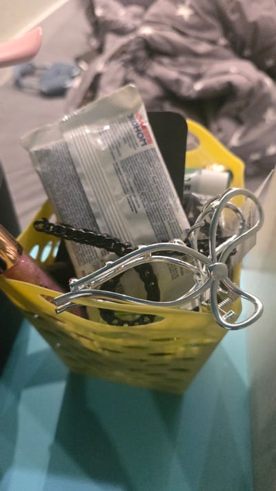 The image shows a close-up of a yellow plastic basket filled with various items. Prominently visible is a silver hair clip shaped like a bow. Behind the hair clip, there is a black chain. To the left of the chain, a tube-like object with text is partially visible, and a pink-colored object is also visible. The basket is sitting on a blue surface, and the background is blurred, showing a grey, textured fabric with white star patterns, possibly a blanket or bedding. The lighting is dim. The image does not provide any information about the city of Ukhta, Russia, or any specific location.