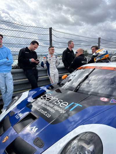 Six adult males are gathered at the Sachsenring motor racing circuit in Hohenstein-Ernstthal, Germany. The foreground features the front portion of a blue, white, and black Porsche race car. The vehicle's hood is adorned with "Sense IT" branding for "Music & Digital Signage for Businesses" and "Melamed Motorsport" logos. An orange and blue racing helmet, bearing a "SIXT" logo, rests on the car's roof near the windshield.
Behind a grey metal barrier, the six males stand. From left to right: one male wears a light blue hoodie and jeans, arms crossed; a second male in dark attire is focused on a smartphone; a third male, identified as a driver by his white and blue racing suit featuring "GP" and "Mobilg" logos, looks forward; a fourth male in dark clothing looks to the right; a fifth male, bald and wearing dark sunglasses with a "GP elite" logo on his dark top, faces left; and a sixth male in dark attire looks to the right. A tall chain-link fence is visible behind the group. The background shows distant structures and natural terrain under a heavily overcast sky. The ground appears dark, consistent with an overcast day at a race track.