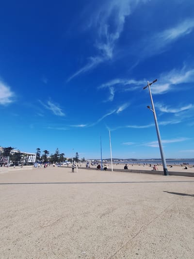 The image depicts a wide, sandy promenade bordering a beach in Essaouira, Morocco. The sky is a vibrant, clear blue with wispy white clouds. On the left side of the promenade, there are buildings and cypress trees. In the mid-ground, a line of people are walking along the promenade and sitting on the beach. A tall, modern lamppost with two lights stands prominently on the right side of the promenade. The beach itself is populated with sunbathers and people enjoying the seaside. The overall scene conveys a sense of a pleasant day at a coastal city.