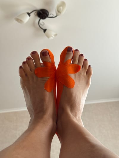 The image shows a close-up, low-angle shot of a person's feet, which are positioned crossed at the ankles, toes pointing upwards. Orange kinesiology tape is applied to the toes of both feet, with the tape on each foot arranged to form a bow-like shape around the big toe. The toenails are painted a dark, glittery red. The background consists of a white ceiling with a dark, multi-headed light fixture and a wall with a textured, light-colored finish. The image does not provide any visual cues to indicate it is located in Vysokaya Gora, Russia.