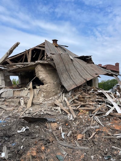 The image shows the remains of a destroyed building in Bilopillia, Ukraine, located at the coordinates 51.138 latitude and 34.286 longitude. The building is a single-story structure with a wooden frame and a metal roof. The roof is partially collapsed, and the walls are heavily damaged. There are pieces of wood and metal debris scattered around the building. The scene appears to be the aftermath of a bombing or other destructive event. The sky is blue with white clouds.