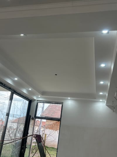 The image displays an interior room in Dolni Tsibar, Bulgaria, depicting an ongoing renovation. The room features white, freshly finished walls and a multi-level white dropped ceiling with a central rectangular recess. At least fifteen illuminated square recessed LED light fixtures are installed along the ceiling's perimeter and stepped sections. A single circular dark opening is present in the central ceiling panel. Exposed electrical wiring extends from the ceiling near the right edge. Large, dark-framed window and door units occupy the left and lower-left portions of the frame, with one section appearing partially open. A smaller, dark-framed window is positioned to the right of the large units. A metallic ladder, draped with red and black hoses or cables, leans against the large window frame in the foreground. Through the windows, an exterior scene reveals residential buildings with red terracotta tiled roofs, bare deciduous trees, a wire fence, and overhead utility lines visible against an overcast sky. No individuals are present in the scene.