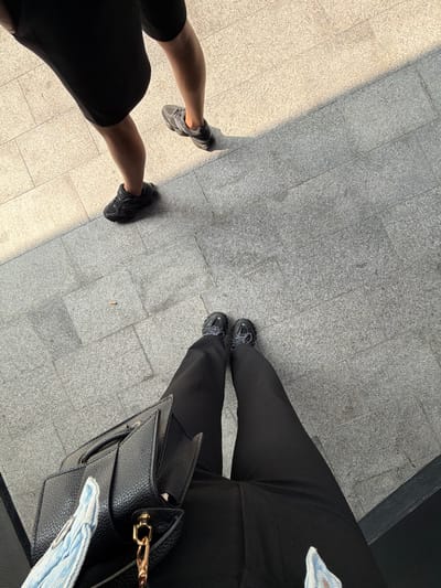 The image shows a view of the ground, revealing two pairs of legs and shoes on a tiled pavement. The tile color varies from light to dark gray due to sunlight and shadow. One pair of legs, on the left, is wearing black shorts or a short skirt and gray sneakers. The other pair is wearing long black pants and dark sneakers, with a black handbag on the right side. The photo appears to be taken in an urban environment, indicated by the tiled pavement. The location is Bayrampaşa, Turkey.