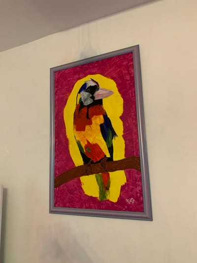 A framed painting displays a colorful bird perched on a branch. The bird has a black, white, and blue head, with vibrant yellow, orange, and green markings on its body. It appears to have a leaf attached to its head. The branch is brown with detailed claws wrapped around it. The background consists of a bright yellow oval shape, surrounded by a field of red and pink. The frame is gray. The painting is mounted on a white wall.