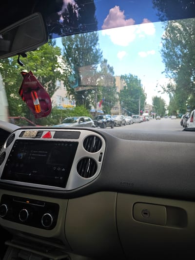 The image is taken from inside a vehicle, looking out through the windshield. A car air freshener shaped like lips with a red and black pattern hangs from the rearview mirror. The dashboard is visible, with the car's infotainment screen displaying song information and playback controls. To the right of the screen are air vents. Below the screen are climate control knobs. The car is on a street lined with trees and parked cars. The sky is blue with white clouds. The scene is consistent with a street in Skadovsk, Ukraine, as indicated by context.
