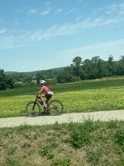 A woman with a helmet is riding a bicycle on a gravel path. She is wearing a pink tank top, black shorts, and a white fanny pack around her waist. The bicycle is a dark reddish-brown color. The path is bordered by dry grass and some green vegetation on the left, and a field of green plants, some of which appear to be sunflowers, on the right. In the background, there are rolling green hills covered in trees and a clear blue sky with some wispy clouds. The scene appears to be a rural or semi-rural area, likely a park or trail system. The location is not specified in the image, and therefore cannot be determined to be in Berg, Austria.