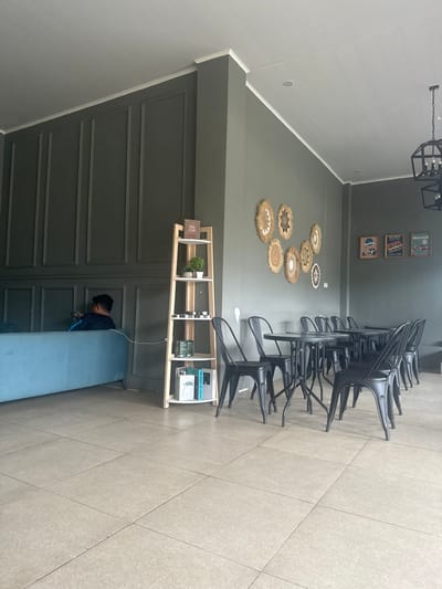 An indoor scene in Sukabumi, Indonesia, depicts a modern and functional space, possibly a cafe or waiting area. The room features a dark gray-green wall with decorative paneling along its lower half, separated by a ledge from a smooth upper section. A lighter gray wall, possibly shiplap, is visible on the left. The floor is covered with light beige/gray tiles.
A black geometric chandelier hangs from the ceiling. Along the dark gray-green wall, a row of several black metal chairs are neatly arranged, possibly stacked. Below these chairs, a light wooden open-shelf unit is positioned. This unit displays a small potted plant, several small canisters, and a stack of books, one clearly titled "THE HEART OF COFFEE." Above the shelf, a cluster of circular, woven wall decorations is mounted. Higher on the wall, three framed pictures, possibly vintage-style posters featuring vehicles, add to the decor.
A light blue upholstered bench runs along the paneled section of the dark gray-green wall. A single person is seated on this bench, facing away from the camera, leaning forward, and looking down. They are wearing a dark shirt. A white cable extends from near the person towards the bookshelf. The overall ambiance suggests a clean, organized, and perhaps quiet environment.
