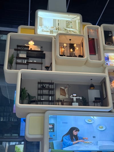 The image displays a large, multi-tiered exhibition structure located in Guangming District, China. This structure consists of numerous stacked and interlocked rounded, rectangular modules, primarily off-white with some yellow accents. These modules serve as either miniature dioramas of interior room designs or digital screens presenting various scenes.

In detail, from top to bottom:
The top-center module is a digital screen showcasing a brightly lit, modern living room and dining area rendering, featuring light-toned furniture.
Below this, to the left, a diorama module presents a room with a tall, dark brown open-shelving unit filled with books and decorative objects against a gray wall. A pink-domed pendant light illuminates the scene, which also includes a small potted plant and a folded chair.
Adjacent to this, a central diorama module features a room with a black chandelier comprising five spherical light sources, an abstract orange-brown artwork on the wall, and the indistinct form of a figure beneath the chandelier.
Further down, a left-side diorama module contains a dark green armchair and another tall, dark brown open-shelving unit with books and decorative items, alongside a potted plant.
To its right, a central diorama module depicts a room with a white humanoid robot holding a tablet, positioned between two dark wooden side tables (one holding a laptop). A multi-sphere chandelier hangs above an abstract artwork, and a dark pendant light is visible. A green plant stands next to the robot.
The lowest visible module is a digital screen displaying a woman with long dark hair, wearing a blue and white striped shirt, seated at a light-colored table and eating with cutlery. The background of this digital scene shows light blue walls, two decorative gold-accented plates, and a television screen. The overall environment appears to be an indoor exhibition space.
