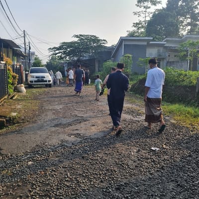 The scene takes place on a gravel road in a residential area in Kecamatan Tamansari, Indonesia.  The road is rough and uneven, with some large potholes.  A white car is parked on the left side of the road, facing the camera. There are nine adults and two children walking on the road. They appear to be walking in the same direction. Some of the adults are wearing traditional clothing. There are houses on both sides of the road. The houses are mostly made of concrete and have flat roofs.  The foliage is lush and green. The sky is blue and clear.  The scene is typical of a residential area in Indonesia.