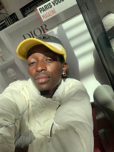 An individual with dark skin, wearing a yellow and grey trucker cap and an off-white puffer jacket with reflective piping, is depicted from the upper chest upwards, directly facing the viewer. A small silver hoop earring is visible in the left ear and a nose piercing on the left nostril. The subject's face and jacket are illuminated by strong, directional sunlight, casting horizontal shadows across the background. The background features a white wall with the large black text "DIOR" prominently displayed. Above and to the right of the "DIOR" logo, a sign reads "PARIS VOGUE" in black and "MAGAZINE" in red. To the far left, indistinct white clothing items are visible on mannequins or displays. A vertical reflective surface, likely a mirror or glass panel, occupies the right side of the frame, showing a partial reflection of the scene. The scene is located inside a commercial or retail space, likely within Paris-Charles de Gaulle Airport, situated in or adjacent to the city of Le Mesnil-Amelot, France.