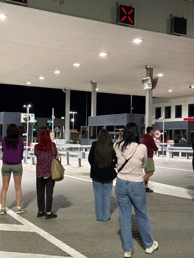 The image depicts a group of five people standing on a paved area, seemingly at a border crossing point. The location appears to be in the Prijepolje Municipality, Serbia, based on the presence of border control infrastructure.

From the top of the image, a man wearing a maroon shirt and khaki shorts stands near a traffic barrier. Below, a woman with long dark hair is wearing a light pink sweater and blue jeans. The next person down is a woman with long dark hair and a black top, also wearing jeans. Further down, a woman with red hair, a patterned purple top, and black trousers is carrying a tan bag. At the bottom of the image, a woman with dark hair and a purple shirt is wearing khaki shorts.

Border control structures are visible, including traffic lights, surveillance cameras, signs with green arrows, and guard posts. The background is dark, suggesting it is nighttime. The lighting is provided by artificial sources illuminating the pavement and buildings. A large, white building with vertical lines is also visible on the left side of the frame. A red 'X' signal light is visible on the building.