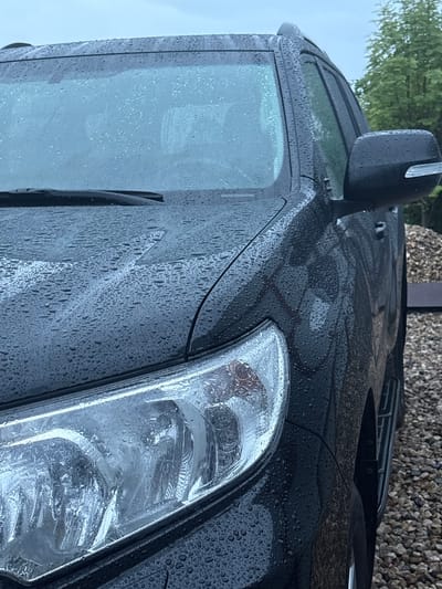 A black SUV is parked outdoors on a cloudy day. The vehicle is covered in water droplets, indicating recent rainfall. The SUV's headlights and side mirror are visible, as is the gravelly ground surrounding it. Trees are visible in the background, suggesting a semi-rural or suburban setting. The image does not provide any specific information about the location in Shebarshino, Russia.