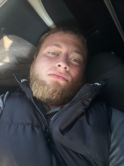 A medium shot selfie of a young man with a beard and blue eyes is taken indoors, possibly inside a vehicle. He is wearing a dark vest over a grey shirt. The background is dark and out of focus, with some blurry shapes that might be seats or fabric. The lighting is somewhat dim, with a natural light source casting a shadow on the left side of his face. The man is looking directly at the camera with a neutral expression. The image does not provide any contextual information about the city of Sculeni, Moldova.