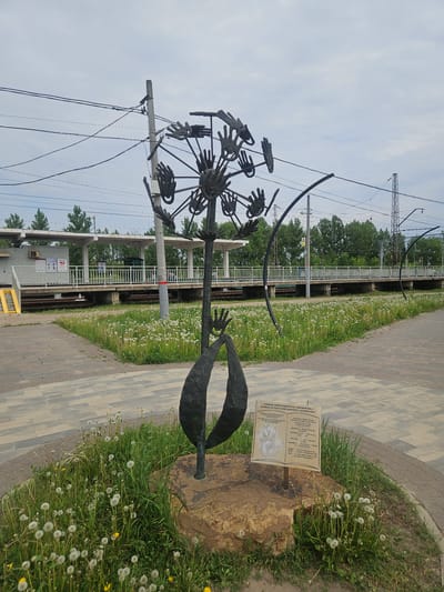 The image shows a metal sculpture in Речицы, Russia, near a railway line. The sculpture is of a dandelion seed head, but instead of seeds, it has hands at the ends of the spokes. The sculpture rests on a rock in a patch of grass dotted with actual dandelions. Beside the sculpture is a small plaque with text in Russian and English, describing the sculpture as a "Symbol of Peace and Kindness." In the background is a railway platform with overhead power lines. The walkway next to the train is made of light gray rectangular pavers. The sky is overcast.