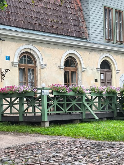 The image shows the exterior of an old building with a weathered facade in Kuldīga, Latvia. The building features arched windows and a tiled roof. A green wooden deck or veranda runs along the front of the building, adorned with planters full of pink and purple flowers. A sign with a blue number "1" is visible on the wall above the first window. A parking sign with a "P" symbol is also present on the deck. The ground in front of the deck is paved with cobblestones, and a patch of green grass is visible on the left side of the image. The overall scene suggests a historical setting, possibly a cafe or restaurant with outdoor seating.