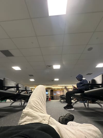 The image depicts an indoor scene, possibly within an airport terminal based on the presence of rows of seating. The perspective is from a seated position, with a person's legs and shoes prominently in the foreground. A second individual in a hooded sweatshirt is seated further away, seemingly engaged with a mobile phone. The background suggests a waiting area, with a view towards the terminal concourse. Specific details that confirm the location as Denver, United States of America, cannot be ascertained from the image alone, as there are no visible landmarks or signage to confirm the location.